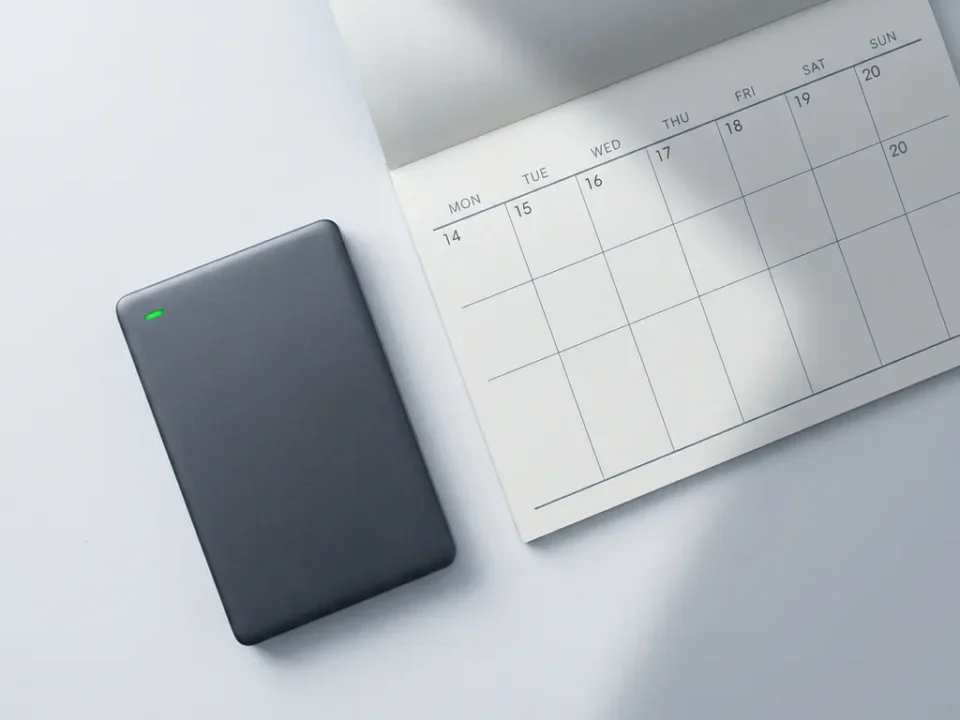 Compacte externe harde schijf op wit bureau naast een open weekkalender, met groen indicatielampje en zachte schaduwen.