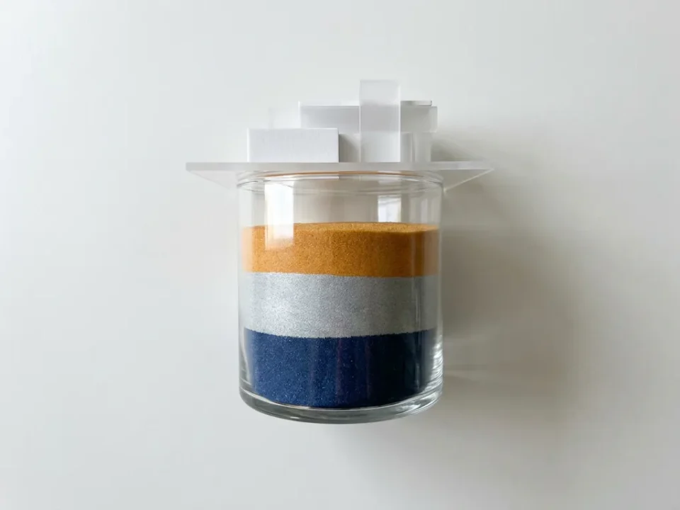 Glazen container met strakke lagen gekleurd zand in marineblauw, zilver en amber, met een architectuurmodel erop, op wit oppervlak.